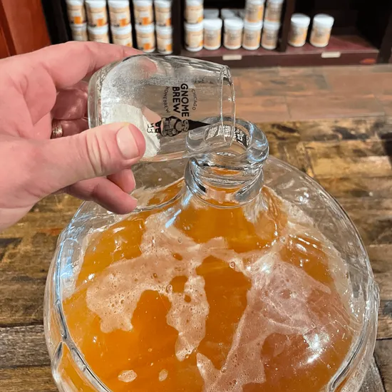 A hand tips pale powder from a Gnome Brew glass into a carboy of amber cider